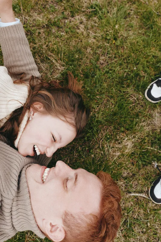Smiling couple lying on grass outdoors, close-up of faces, cozy sweaters, warm natural light, relaxed mood, affection and connection, candid lifestyle moment with joyful energy.