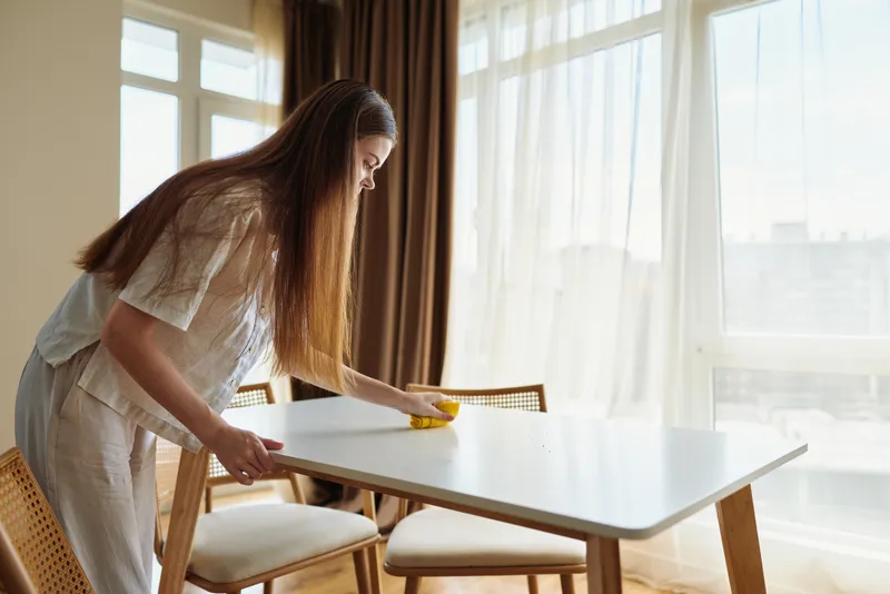 Woman cleaning a dining table with a yellow sponge in a bright home interior, wiping the wooden surface near sunlit windows, casual morning chores and domestic tidying scene.