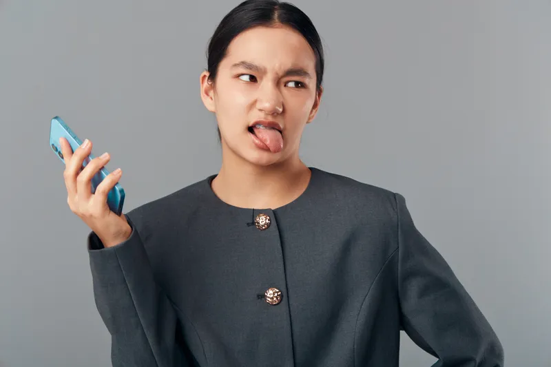 Woman holding smartphone with displeased face and sticking out tongue. Adult female in gray jacket showing annoyance or irritation on gray background in studio portrait.