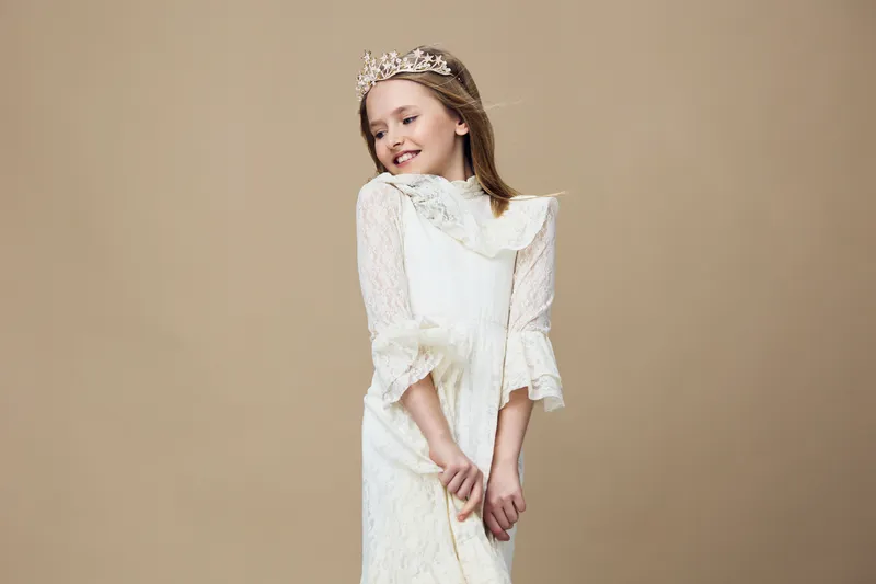 A young girl wearing a beautiful white dress and a crown, showcasing joy and innocence against a soft beige background, perfect for celebrating childhood moments