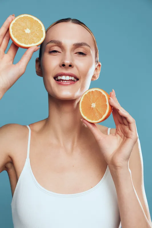 Smiling woman holding two halves of fresh orange near her face. Close-up portrait with clear skin and natural makeup on blue background. Concept of healthy lifestyle and freshness.