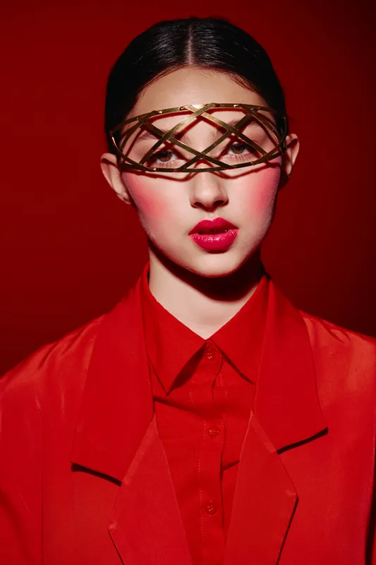 Creative fashion portrait of a woman with glossy makeup and a leather headpiece. Red outfit and background create a bold, high-contrast beauty editorial concept in studio lighting.