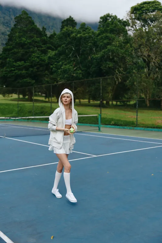 Young woman in sporty white outfit holding a tennis ball on a court, showcasing athleticism and confidence in a vibrant green outdoor setting