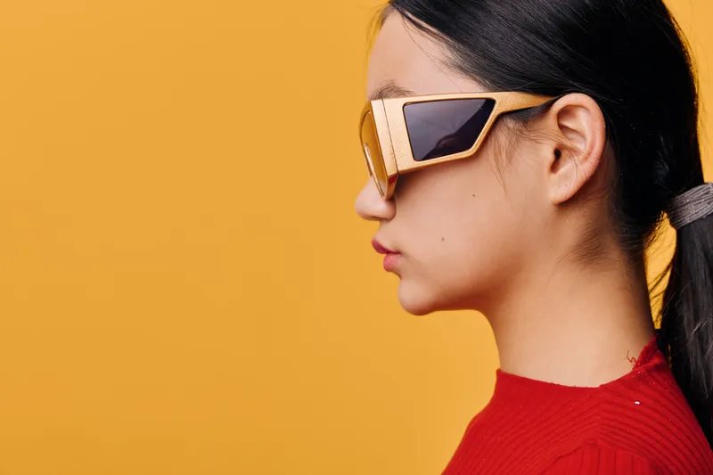 Profile of young woman wearing wooden sunglasses and red sweater against yellow background in studio. Fashion, style and beauty concept with clean minimalism and modern accessories in portrait.