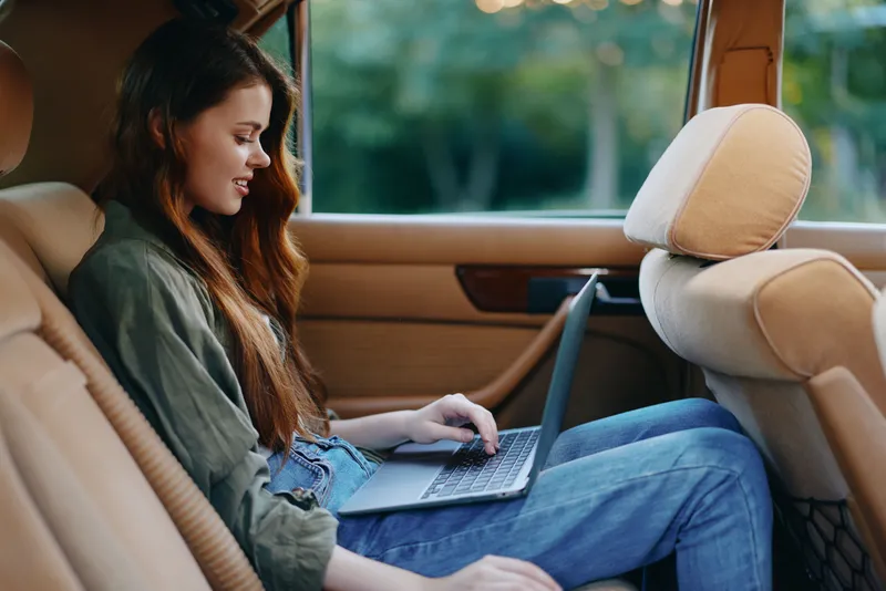 Young woman working on a laptop in the backseat of a car, showcasing a relaxed and focused expression while dressed casually, with a soft green background.