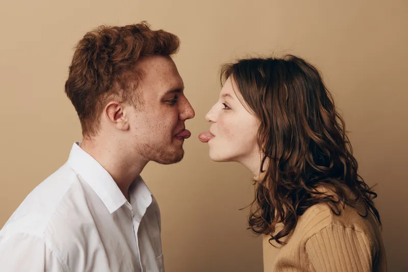 Young man and woman facing each other playfully sticking out tongues. Casual clothing and neutral background create a fun and lighthearted mood of friendship and humor.