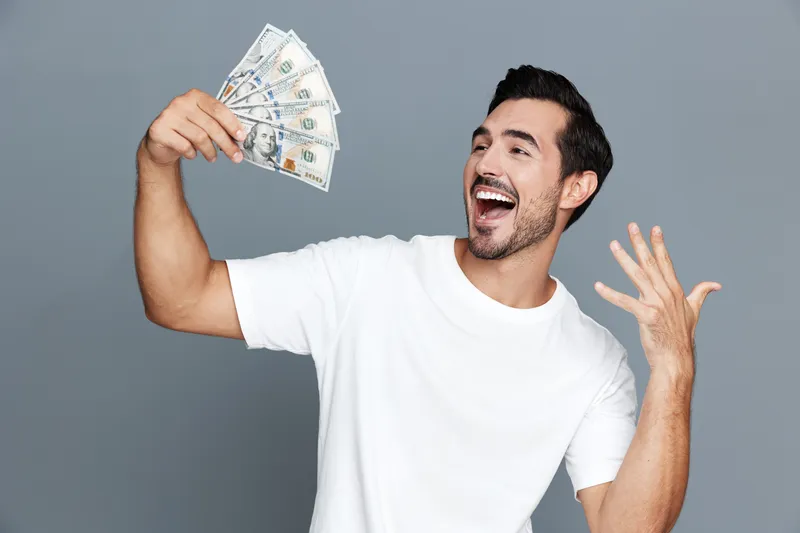 Joyful man celebrating success, holding cash in hands, smiling broadly with excitement. Isolated on plain gray background, studio portrait, lifestyle concept.
