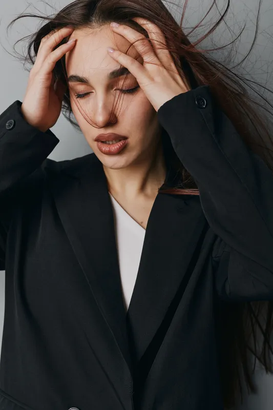 Woman with long hair and closed eyes holding head with hands, feeling stressed or tired. Portrait of young female in black jacket with natural makeup on gray background.