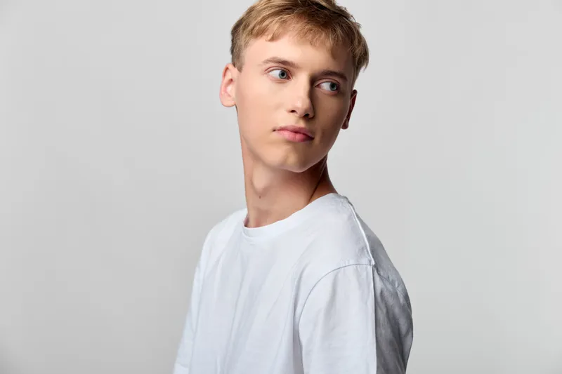 Young man in a white t shirt stands against a neutral gray background, looking to the left with a pensive expression, clean studio composition and minimal detail