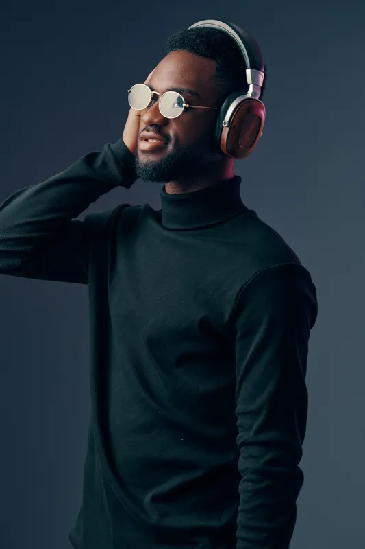 Young man enjoying music with headphones, wearing stylish glasses, and a black turtleneck sweater against a dark background, exuding a relaxed vibe