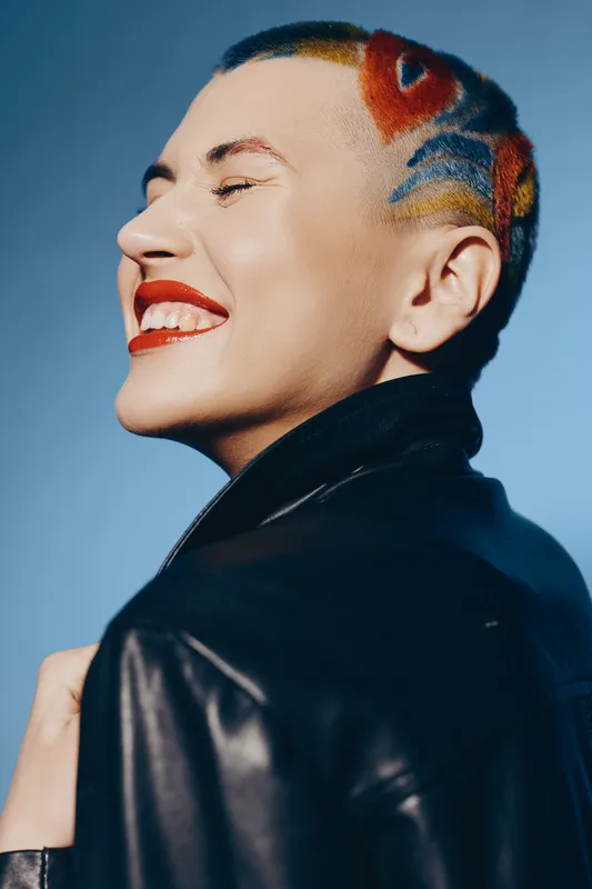 Smiling woman with colorful short hair and vibrant makeup wearing a black leather jacket against a soft blue background, expressing joy and individuality.