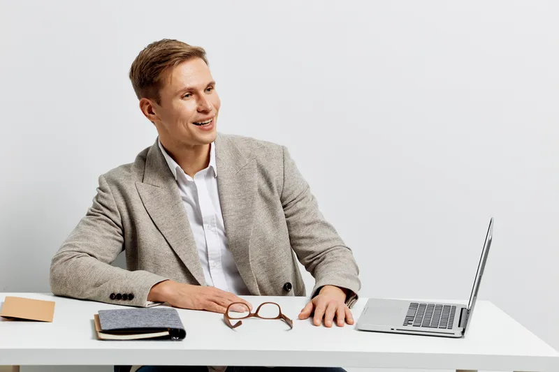 Professional young man working at a modern desk with laptop, glasses, notebook, and smartphone. Happy and focused, dressed in smart casual, neutral background. office workspace concept