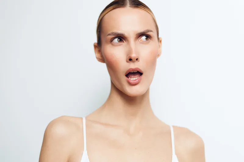 Surprised woman with open mouth looking sideways, neutral background, natural skin tone, minimal makeup, wearing white top, expressive facial expression, studio lighting.