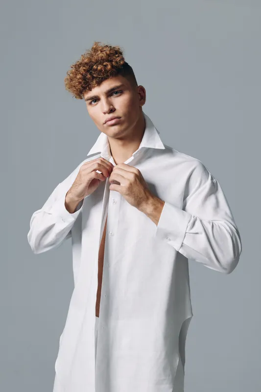 Young man with curly hair wearing a white shirt unbuttoning collar against gray background with neutral expression standing studio portrait of confident fashion model casual style.