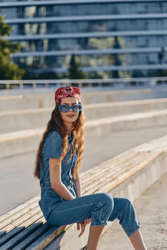 lifestyle woman in modern boho-western denim street style wearing sunglasses and a red bandana with film color effect sitting outdoors in urban setting