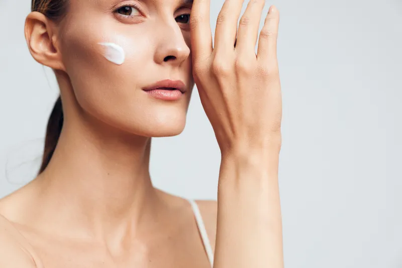 Woman applies facial cream on face with fingers near cheek, showing skincare routine and personal hygiene. Bright studio light highlights smooth skin, beauty care for self care.