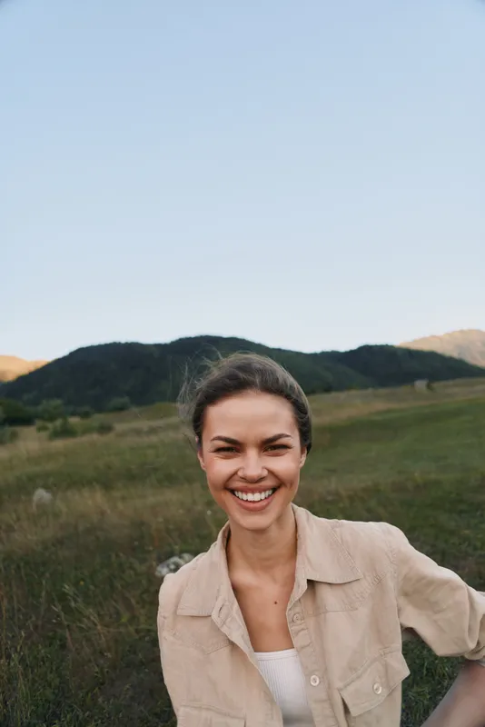 A smiling young woman wearing a light beige shirt stands in a sunlit grassy hillside field, creating a candid outdoor lifestyle portrait with relaxed confidence.