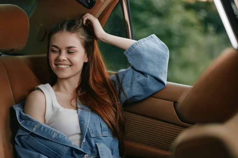 Smiling young woman in casual denim jacket enjoying a moment in a car, with vibrant greenery in the background, showcasing carefree emotions and summer vibes.