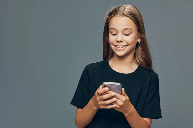 Girl with long hair wearing black t shirt smiling and looking at smartphone in her hands on grey background. Portrait of child using mobile phone and enjoying digital content.