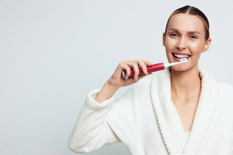 Young woman in white bathrobe brushes teeth with electric toothbrush in bathroom smiling showing dental hygiene routine and healthy oral care isolated on light background.