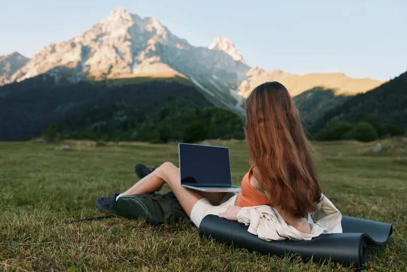 A person with long hair sits on a mat in a grassy field, using a laptop outdoors. The scene captures solitude, focus, and a calm, productive work vibe against a mountain landscape.
