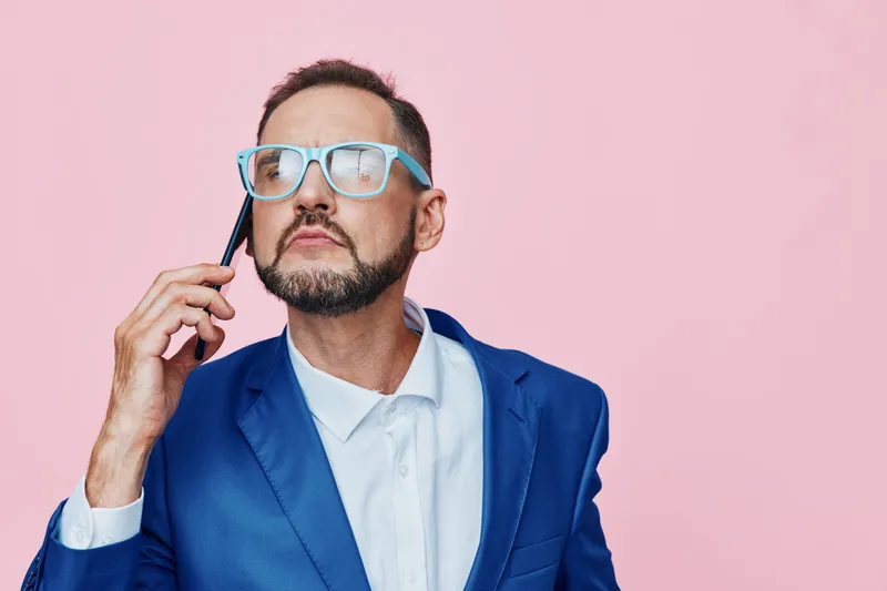 A professional businessman stands against a solid pink background, wearing a blue suit and glasses, talking on a phone with a thoughtful expression, conveying focus and calm authority.