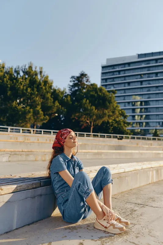 Lifestyle woman in modern boho-western denim street style sitting relaxed outdoors, enjoying sunny day, film color tones with casual chic outfit and red bandana.