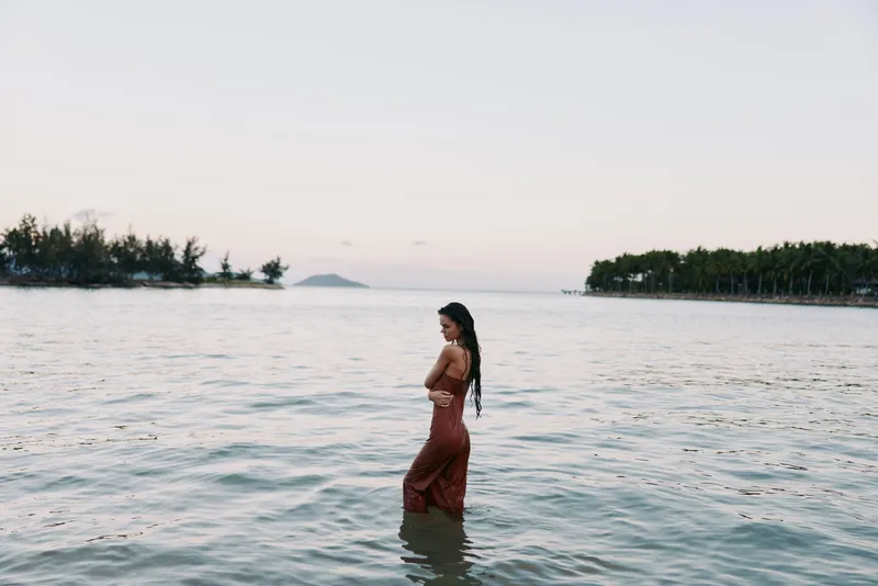 A woman stands waist-deep in calm tropical water, wearing a red dress with a long braid, as soft light kisses the lagoon and distant islands.