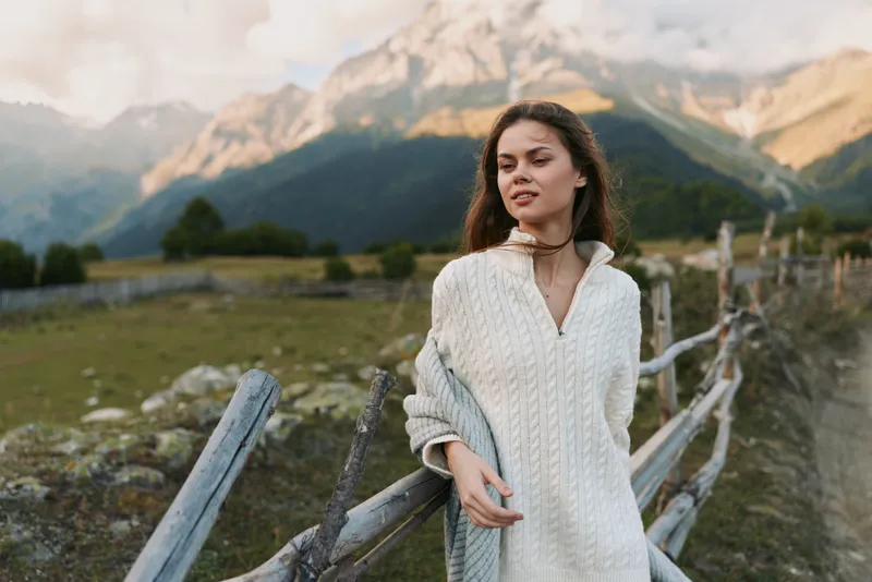 Young woman in a white knit sweater stands by a rustic wooden fence with a dramatic mountain landscape behind, conveying calm and outdoor lifestyle.
