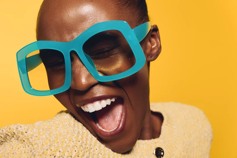 Close-up of a joyful black woman with dark skin wearing oversized blue glasses and a beige sweater, smiling widely against a bright yellow background. Concept of happiness and fashion.