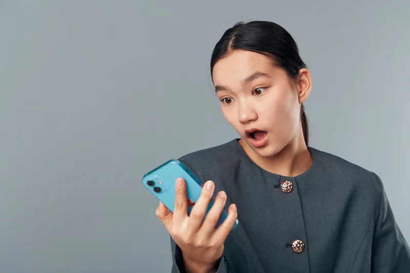 Young woman with surprised expression looking at smartphone with open mouth on gray background. Female person wearing dark jacket reacting to unexpected message or notification on phone.