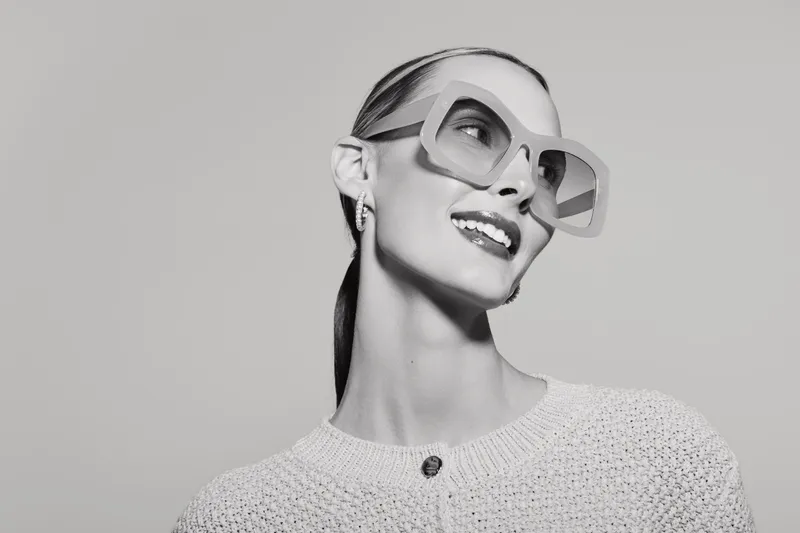 Stylish young white woman with pale skin wearing oversized glasses and a knitted sweater smiles confidently against plain background in black and white portrait.