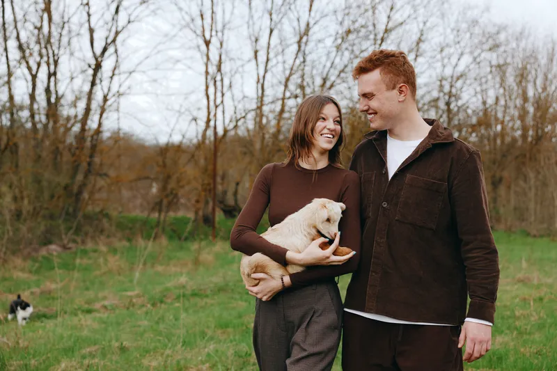 A joyful couple outdoors with a small goat kid cradled by the woman, while the man smiles nearby. A relaxed lifestyle moment highlighting companionship, rural charm, and gentle animal interaction in