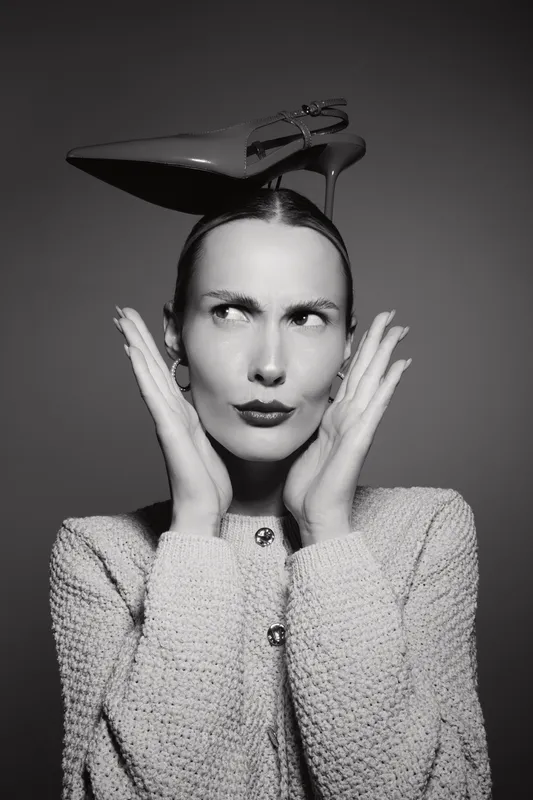 Caucasian woman with light skin wears high heel shoe on head and looks sideways with hands near face in black and white studio portrait.
