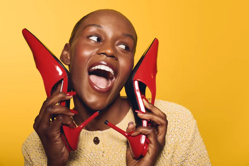 Joyful dark skinned African woman with shaved head holds red high heels near face, smiling widely on yellow background. Concept for fashion, happiness, style, and beauty expression.
