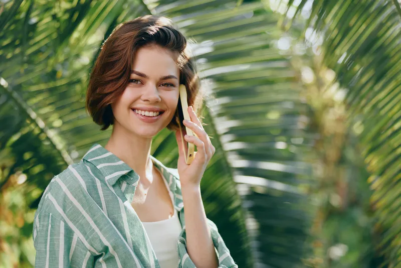 A smiling woman stands outdoors, talking on a smartphone, with lush palm leaves in the background and warm sunlight highlighting her relaxed pose.