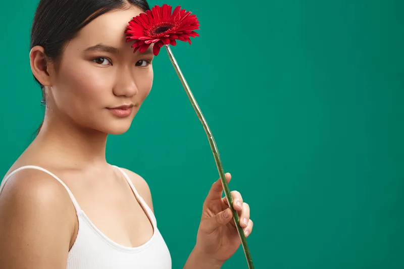 Asian woman stands against a solid green backdrop, wearing a white tank top and holding a red flower near her face, creating a calm, graceful portrait with natural poise.