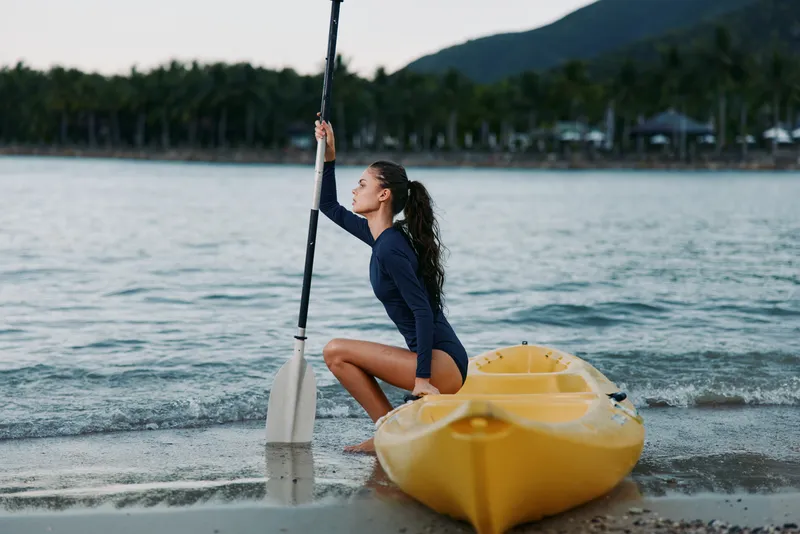 A young woman kneels at the shoreline beside a yellow kayak, gripping a paddle as gentle waves lap the sand under soft natural light. She wears a navy swimsuit, hair tied back, conveying calm outdoor