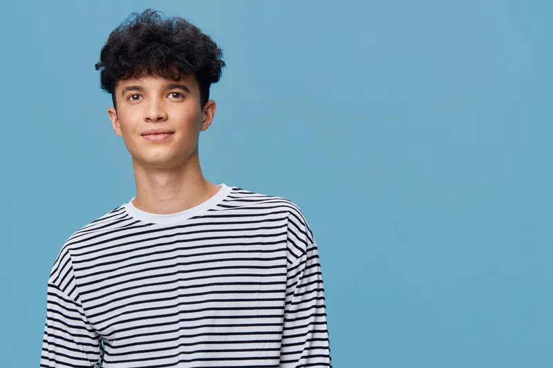 Young man wearing casual striped shirt with curly hair standing against blue background, smiling slightly and looking confident in a bright studio setting.