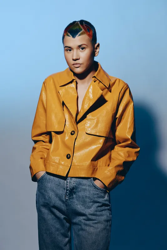 Young woman with colorful short hair wearing a shiny mustard jacket and relaxed denim pants against a soft blue background, styled in modern casual fashion.