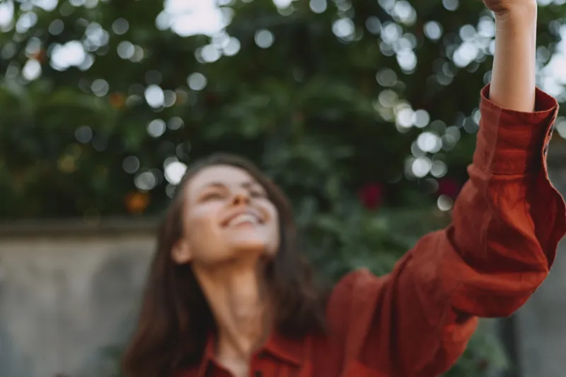 A joyful woman in a red jacket raises her arm and smiles outdoors, capturing a carefree lifestyle moment. A bright, candid concept focused on happiness, spontaneity, and connection with nature