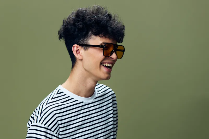 smiling man wearing vintage sunglasses and striped shirt with curly hair on olive background, joyful young adult male portrait in casual style, bright lighting.