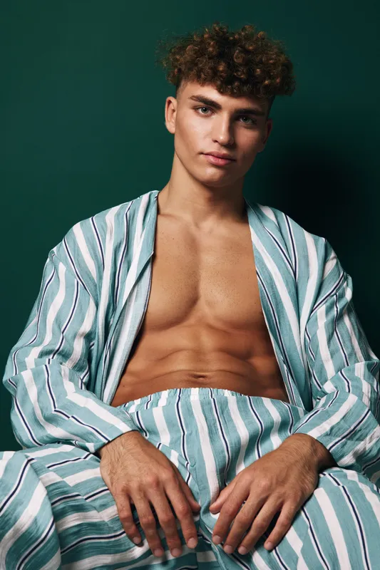 Young man with curly hair wearing striped open shirt and pants sitting relaxed against dark green background with confident expression and casual pose.