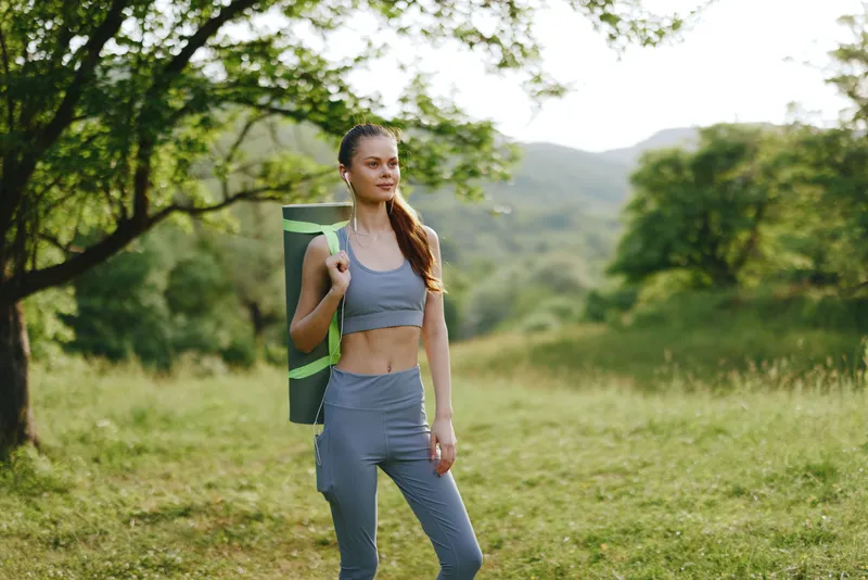 Young woman in athletic wear carrying yoga mat in nature, showcasing fitness, healthy lifestyle, and outdoor activity during summer