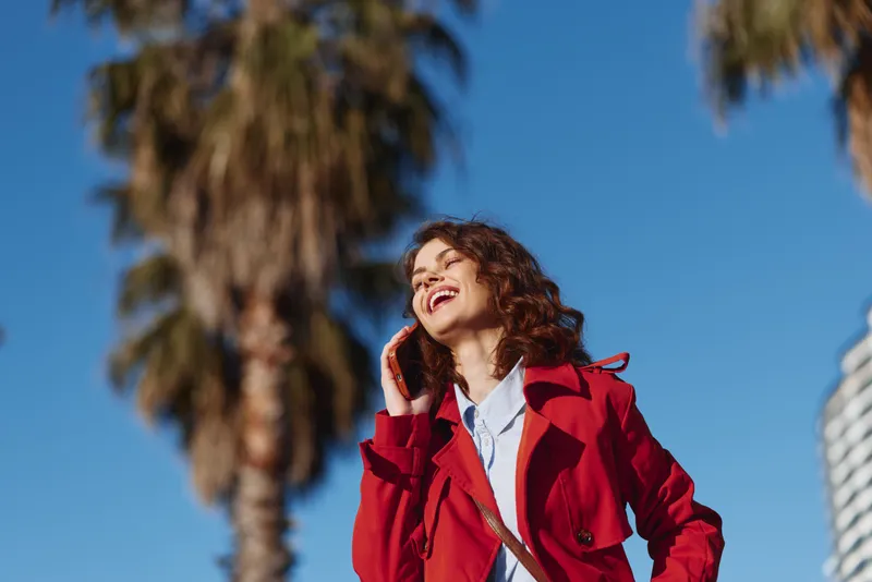 Smiling woman wearing red coat poses outdoors under blue sky with palm trees, bright sunny mood, fashion portrait with bold colorful styling and casual confidence on a sunny urban street