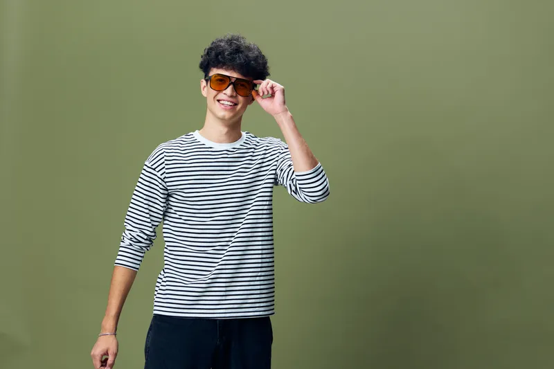 Smiling young man wearing striped shirt and orange sunglasses with curly hair standing against olive green background, casual style, studio portrait.