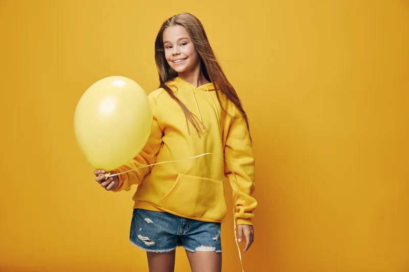 Girl with long hair wearing yellow hoodie and denim shorts holding yellow balloon isolated on yellow background. Smiling child standing with casual outfit and playful mood.