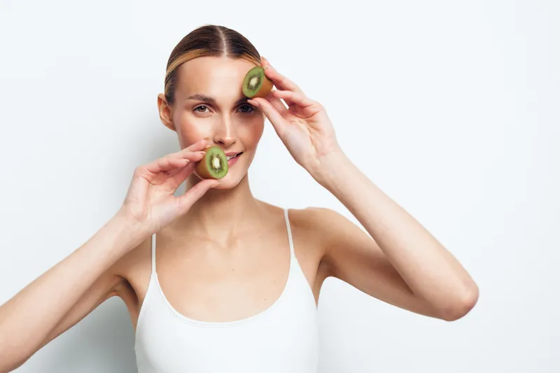 Young woman holding fresh kiwi slices near her face smiling, clean skin, healthy lifestyle, white background, natural beauty, casual outfit, bright lighting