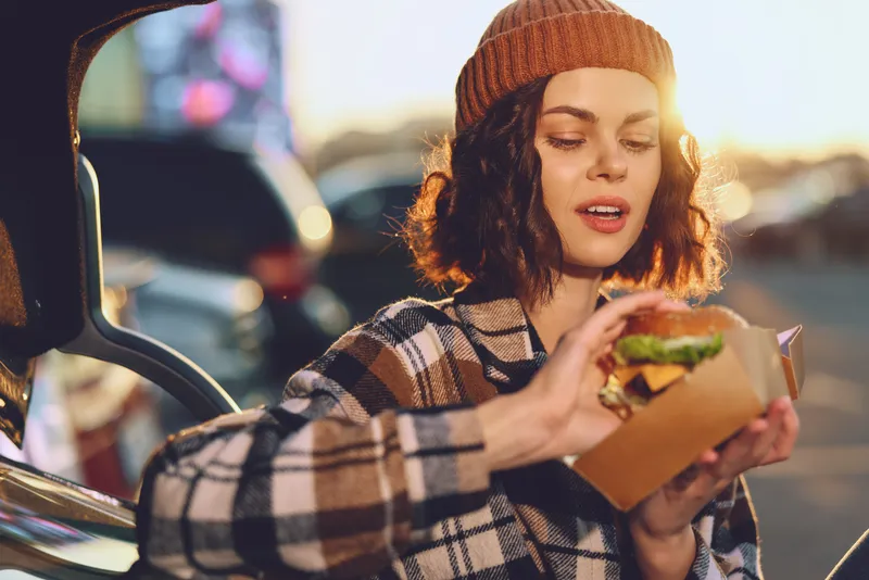 Woman in beanie holding burger by car trunk at sunset, candid urban portrait with authenticity and golden hour glow, emotional storytelling of mindful living and cozy modern lifestyle.