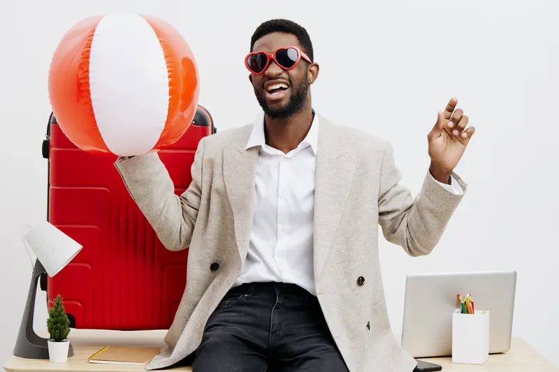 Cheerful young man with dark skin, stylish beige blazer, and red sunglasses, holding a large beach ball while smiling and looking joyful. He is sitting at a desk with a red suitcase, small potted plan